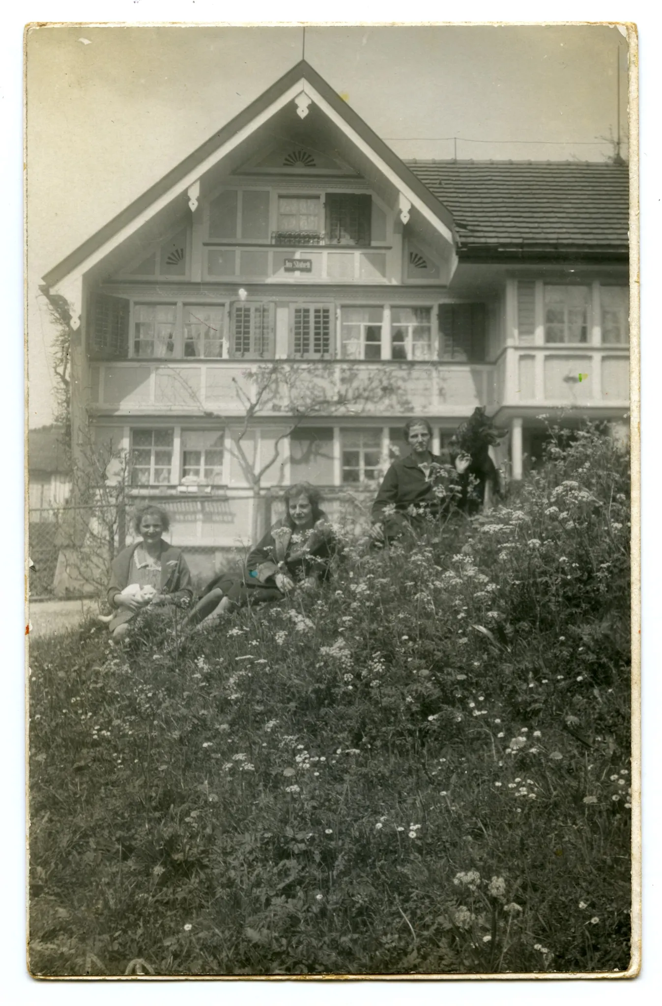 Ansicht mit Hausbewohnerinnen (?) sitzend auf der Wiese vor dem Haus