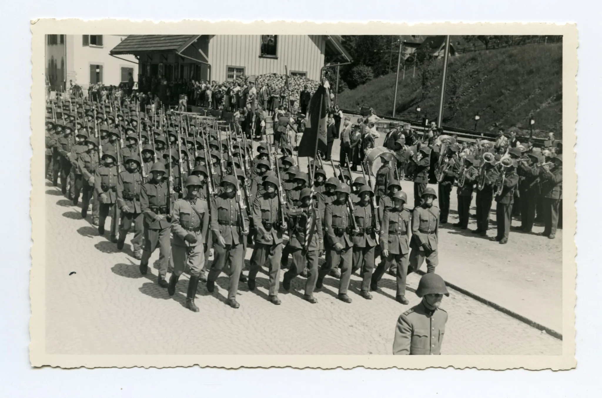 Soldaten (Kompanie) marschieren auf der Strasse (Ebni) Richtung Zeughaus, daneben Militärkapelle vor den Bahnschienen
