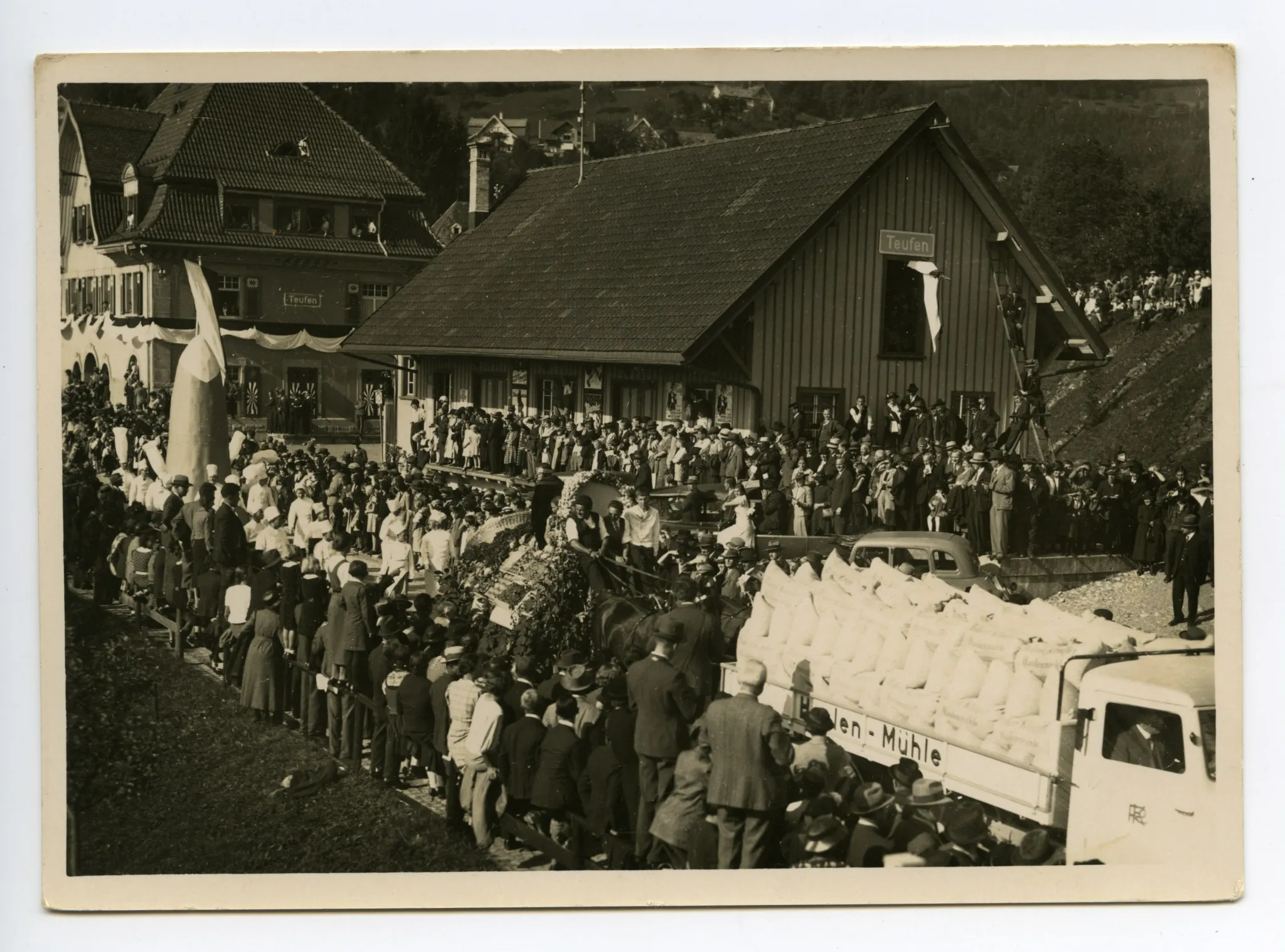Lastwagen beladen mit Mehlsäcken der Haslen-Mühle?, gefolgt von Fuhrwerk der Obst- und Gemüsebauern, gefolgt von Konditoren auf der Strasse, Ebni, Güterschuppen und Bahnhof sichtbar