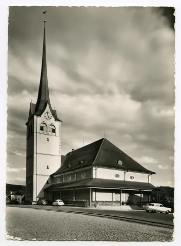 vom Vorplatz des alten Bahnhofs gesehen, mit Bahngeleise. Drei parkierte Autos, davon 2 VW-Käfer entlang der Kirche