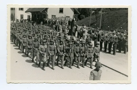 Soldaten (Kompanie) marschieren auf der Strasse (Ebni) Richtung Zeughaus, daneben Militärkapelle vor den Bahnschienen