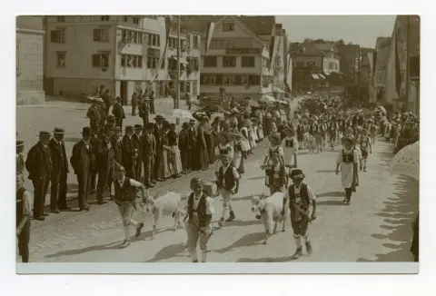 Knaben in Sennentracht und Ziegen, sowie Mädchen in Tracht laufen vor der Häuserzeile mit Gasthaus Anker, Gasthaus zum Adler und Villa Roth, Zuschauer stehen auf der Bahnschiene