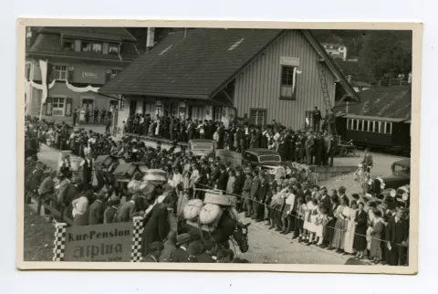 Saumpferde beladen mit Sennengeschirr gefolgt von Sennen mit Tanse auf dem Rücken vor dem Güterschuppen